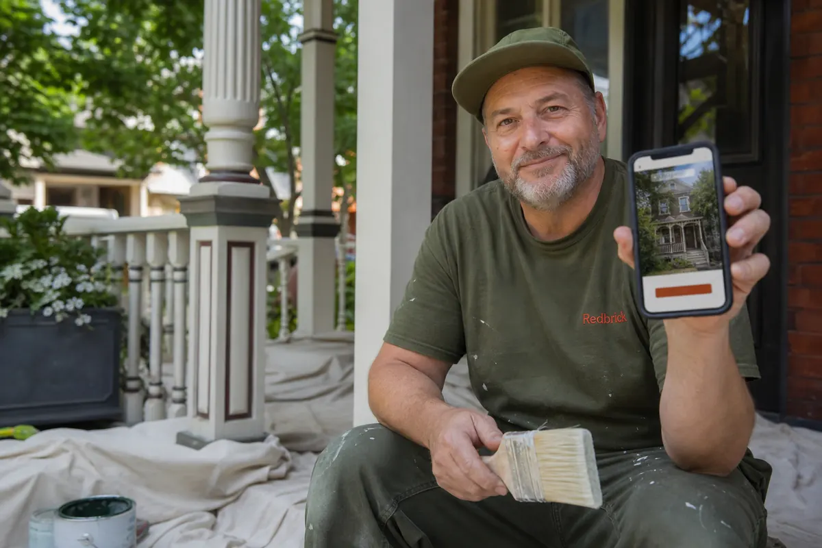 House painter on a front porch of a Roncesvalles Victorian, brush in hand, checking a new quote request on his phone
