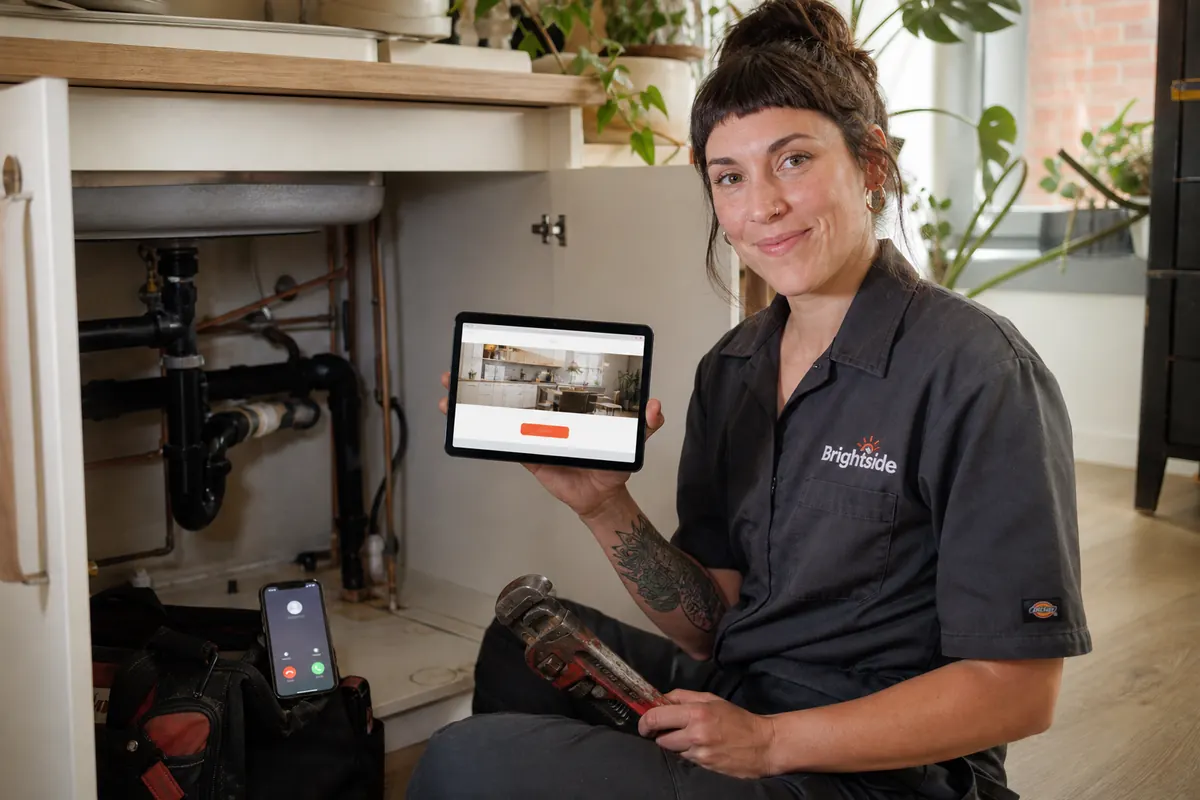 Female plumber under a kitchen sink in a Riverdale semi with her phone beside her showing a customer's call